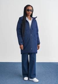 Parka blu navy con cappuccio, chiusura con zip, tasche multiple e polsini regolabili. Abbinata a jeans di denim larghi e sneakers bianche.