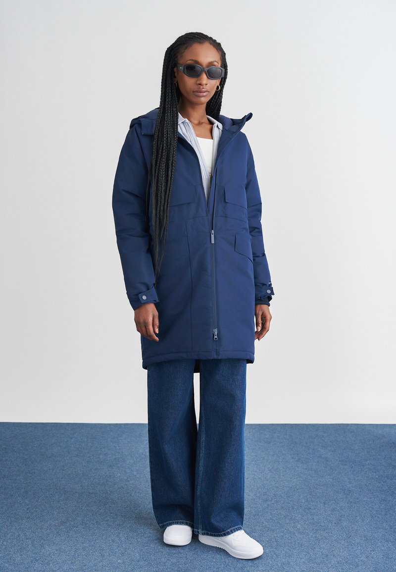 Parka blu navy con cappuccio, chiusura con zip, tasche multiple e polsini regolabili. Abbinata a jeans di denim larghi e sneakers bianche.