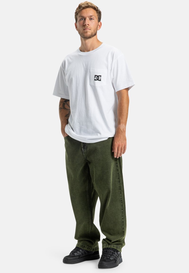 Biała bawełniana koszulka z kieszenią na piersi, z czarnym logo, w połączeniu z luźnymi, ciemnozielonymi dżinsami i czarnymi trampkami.