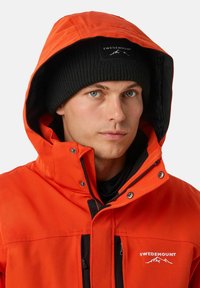 Giacca impermeabile arancione con fodera interna nera, tasche con zip e un logo distintivo. Un berretto lavorato a maglia nero completa il look.