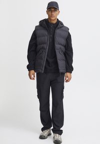 Črna podložena brezrokavnik s kapuco, oblečen nad črno dolgimi rokavi, v kombinaciji s črnimi cargo hlačami in sivimi čevlji. Minimalistična zasnova, preprosta strojna oprema.