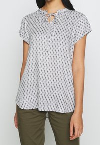 White blouse with black floral pattern, round neckline, short sleeves, and a tie closure at the front. Soft, lightweight fabric.