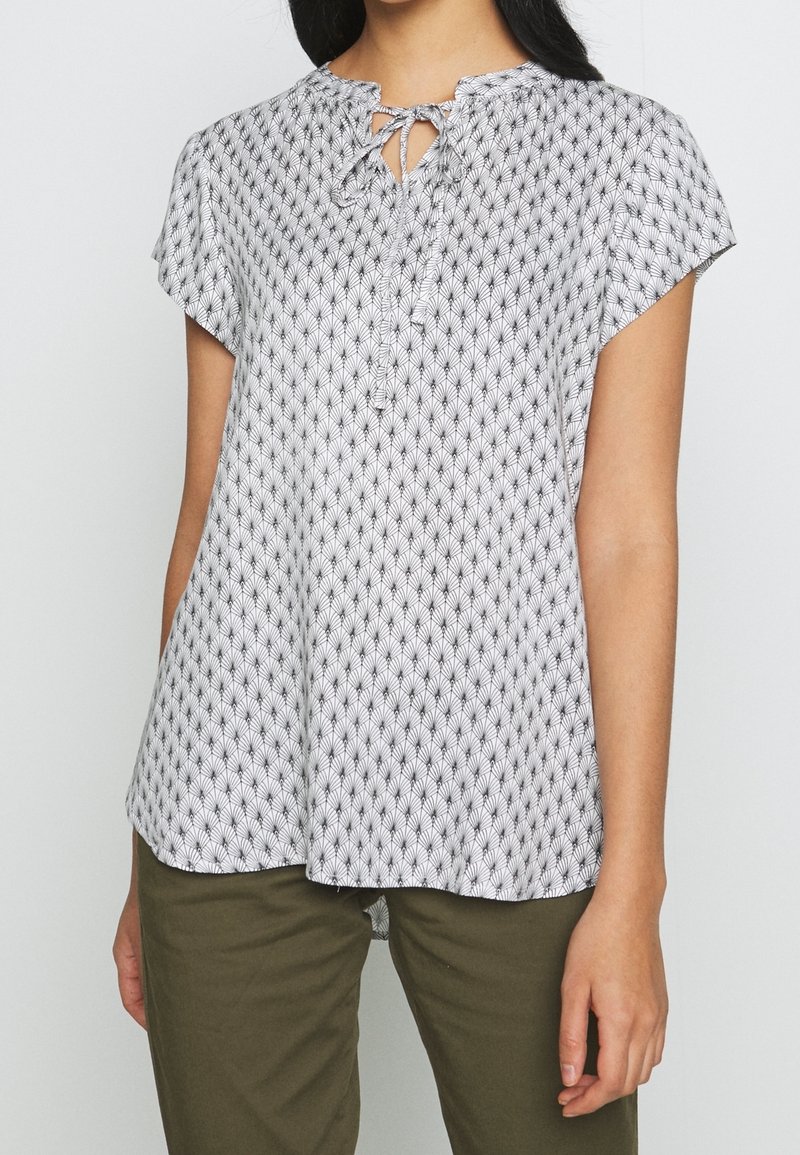 White blouse with black floral pattern, round neckline, short sleeves, and a tie closure at the front. Soft, lightweight fabric.