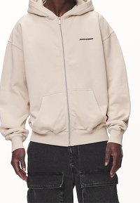 Beige zip-up-hoodie i mjukt tyg, med en känguruficka och ribbade muddar. "PEGADOR"-logotyp tryckt i svart på bröstet.