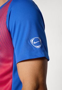 Blå atletisk t-shirt med rødt diagonalt mønster, glat tekstur, og Nike-logo broderet på venstre ærme. Korte ærmer, tætsiddende design.
