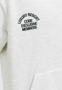 Jasnoszara bluza z miękką teksturą; na piersi ma czarne haftowane logo z napisem "LUXURY RESORT CORE EXCLUSIVE MEMBERS".