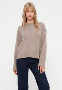 Beige pletený svetr s ribbovanými lemy a manžetami, s uvolněným střihem a kulatým výstřihem. V kombinaci s tmavě modrými džínami s širokými nohavicemi.