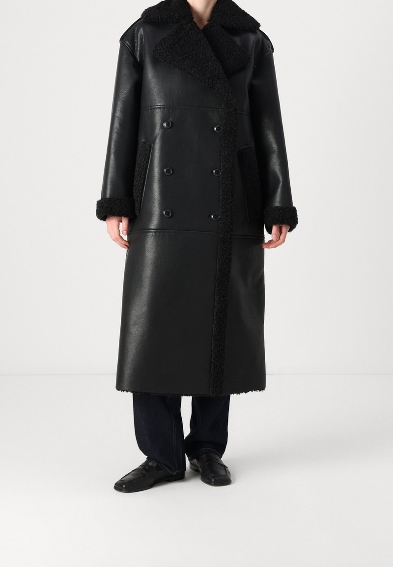 Personne portant un long manteau noir en cuir à double boutonnage, avec des finitions en peau lainée au col, aux poignets et sur les poches, associé à un pantalon foncé et des chaussures noires.