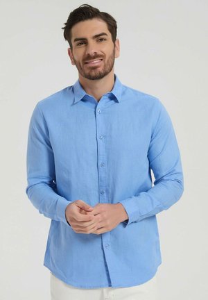 Homme à la barbe taillée portant une chemise bleu clair à manches longues boutonnée, debout devant un fond clair uni, les mains jointes.