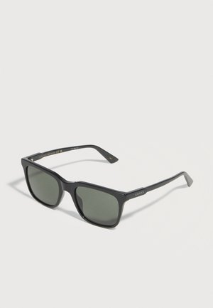 Lunettes de soleil Gucci rectangulaires noires avec verres teintés foncés et logo Gucci visible sur les branches, posées sur une surface blanche.