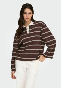 Pruun pika varrukaga polomees valgete horisontaalsete triipudega, vööpikksärk kaelusega, kaelusel kolm nööpi ja lõdva lõikega, sobitatud heledate pükstega.