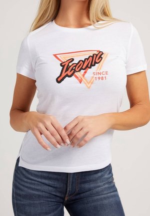 Kobieta w białej koszulce z krótkim rękawem z napisem "Iconic Since 1981" oraz pomarańczowym trójkątnym wzorem, w połączeniu z ciemnoniebieskimi dżinsami.