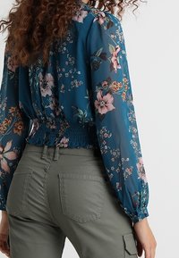 Blomstret bluse i teal med lange ærmer og smocket talje; har pink og hvide blomsterdesigns. Sammen med olivengrønne bukser.