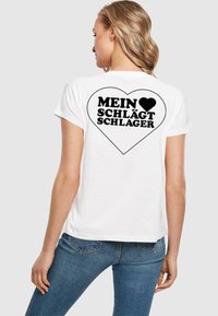 Bílý bavlněný tričko s grafickým designem černého srdce a textem "MEIN ♥ SCHLÄGT SCHLAGER" uvnitř obrysu srdce na zadní straně.