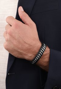 Zwarte gevlochten leren armband met zilverkleurige accenten, gedragen om een pols. Heeft een gestructureerd ontwerp en een strakke pasvorm tegen de huid.