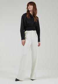 Chaqueta negra corta con cuello alto, combinada con pantalones de pierna ancha blancos con detalles en crema y una textura suave. Tacones puntiagudos negros.