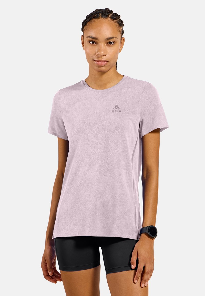 Junge Frau mit geflochtenem Haar trägt ein helllila kurzärmeliges Shirt und schwarze Shorts und steht vor einem schlichten hellen Hintergrund.