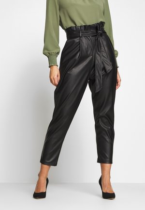 Femme portant un pantalon en cuir noir taille haute avec une ceinture nouée, associé à un chemisier vert à manches longues et des talons noirs.