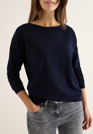 Maglione blu navy con scollo ampio e maniche a tre quarti, realizzato in tessuto morbido, abbinato a jeans grigi con una texture sbiadita.