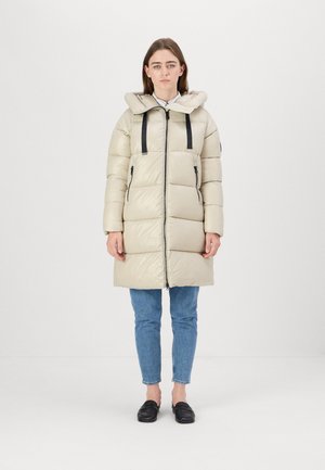 Lichtbeige pufferjack met een capuchon, voorzien van een ritssluiting aan de voorkant, zwarte banden en zijzakken. Gekozen in combinatie met blauwe jeans en zwarte schoenen.