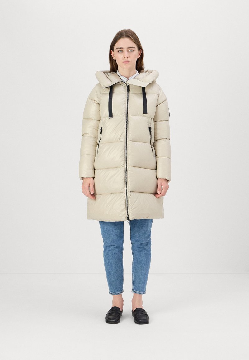 Light beige puffer coat with a hood, featuring a front zipper, black straps, and side pockets. Worn over blue jeans and black shoes.