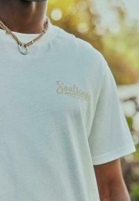 T-shirt en coton blanc avec un logo "Soulscape" doré sur la poitrine. Manches courtes et texture douce. Porté avec un collier en corde marron.