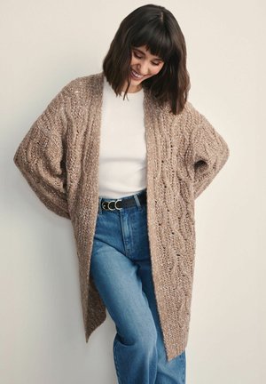 Długi, oversize kardigan z fakturowanej beżowej włóczki z wzorem w warkocz. Noszony na otwarto na białej bluzce, w zestawieniu z niebieskimi dżinsami z wysokim stanem i czarnym paskiem.