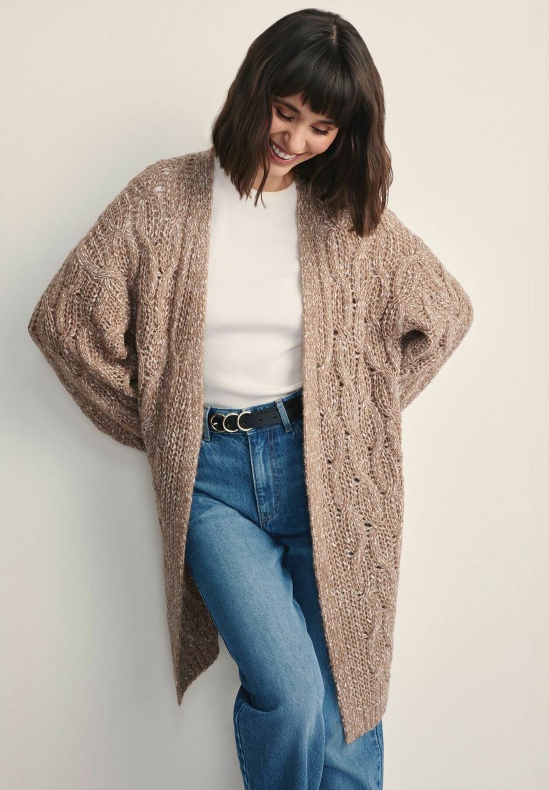 Długi, oversize kardigan z fakturowanej beżowej włóczki z wzorem w warkocz. Noszony na otwarto na białej bluzce, w zestawieniu z niebieskimi dżinsami z wysokim stanem i czarnym paskiem.