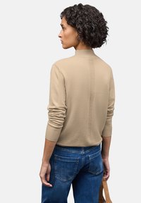 Beige pletený svetr s vysokým límcem, žebrovanými manžetami a švem na zadní straně, v kombinaci s modrými džíny a béžovou kabelkou.