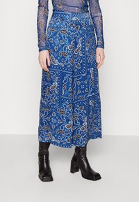 Žena v modré midi sukni s paisley vzorem, černých vysokých botách a průhledném topu s dlouhým rukávem, který má odpovídající paisley design.