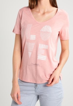 Kobieta ubrana w różowy T-shirt z dekoltem w serek z abstrakcyjnym białym napisem "LOVE" oraz jasnoszare spodnie, stojąca na jednolitym tle.