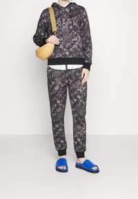 Felpa nera mimetica con cordini, pantaloni coordinati e sandali blu; presenta anche una borsa a spalla color senape.