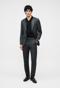 Άνδρας με σγουρά μαλλιά, ντυμένος με σκοτεινό γκρι κοστούμι, μαύρο πόλο πουκάμισο και μαύρα παπούτσια, στέκεται με το ένα χέρι στην τσέπη.