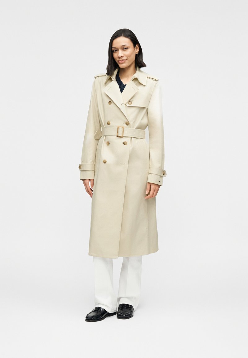 Γυναίκα φοράει μπεζ trench coat με ζώνη πάνω από λευκό παντελόνι και μαύρα loafers, στέκεται σε απλό φωτεινό φόντο.