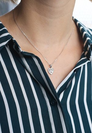 Zilveren hartketting met uitsnijdingen, gedragen op een gestreepte groen-witte blouse met kraag. De ketting is delicaat en van gemiddelde lengte.