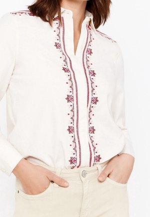 Vrouw draagt een wit blouseje met knopen en rode en zwarte geometrische borduursels over de voorkant en schouders, handen in de lichte beige broekzakken.
