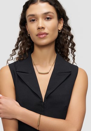 Black sleeveless top with a collar, paired with a gold bar necklace featuring a textured design and a thin gold bracelet on the wrist.
