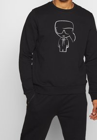 Czarna bluza z bawełnianej mieszanki z białą grafiką kreskówkową przedstawiającą postać w okularach przeciwsłonecznych. Ściągacze na rękawach i u dołu.