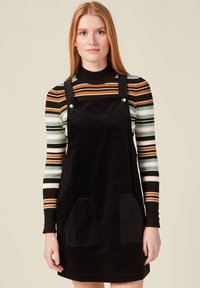 Mustat corduroy-mekko, jossa on säädettävät hihnat, etutaskut ja hopeanväriset nepparit. Vaatteena päällekkäin raidallinen pitkähihainen paita.