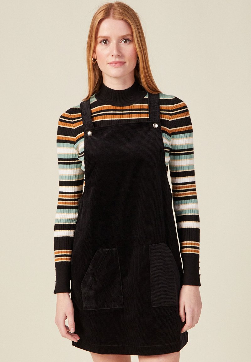 Mustat corduroy-mekko, jossa on säädettävät hihnat, etutaskut ja hopeanväriset nepparit. Vaatteena päällekkäin raidallinen pitkähihainen paita.