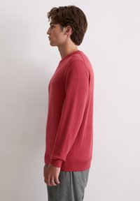 Czerwony sweter dziergany z miękkiej tkaniny, z okrągłym dekoltem i ściągaczami na rękawach, w zestawieniu z szarymi spodniami. Widok z boku.