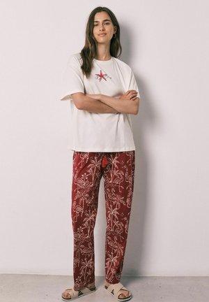Vrouw staat met gekruiste armen, draagt een wit t-shirt met rood zeesterrenontwerp, rode broek met patroon en beige sandalen tegen een effen muur.