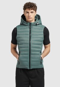 Piepildīts veste teiks zaļā krāsā ar kapuci, priekšējo rāvējslēdzēju un sānu kabatām. Izgatavota no viegla materiāla ar horizontālu quilted dizainu.