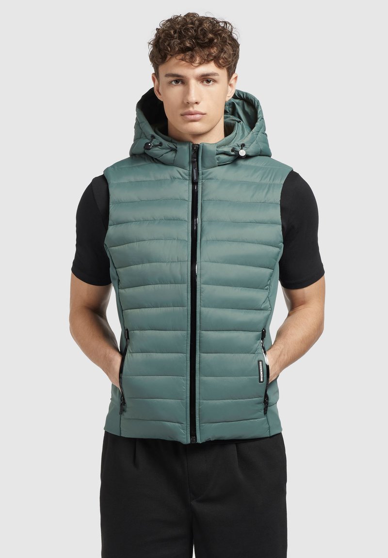 Piepildīts veste teiks zaļā krāsā ar kapuci, priekšējo rāvējslēdzēju un sānu kabatām. Izgatavota no viegla materiāla ar horizontālu quilted dizainu.