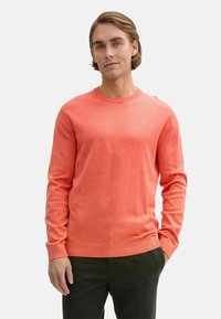 Pulover coral cu guler rotund, confecționat dintr-un material ușor. Prezintă mâneci lungi și mansete din tricot, asortat cu pantaloni închis la culoare. Textură netedă.