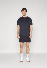 Herre treningsantrekk med mørk marineblå kortermet skjorte og matchende shorts. Skjorten har en logo, og modellen har på seg hvite joggesko.