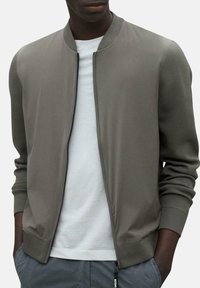 Chaqueta bomber gris con cuello y puños acanalados, con una textura suave y cierre de cremallera, llevada sobre una camisa blanca.
