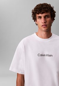 Hvit T-skjorte i bomull med korte ermer, med "Calvin Klein"-logoen i svart over brystet. Mykt, avslappet design.