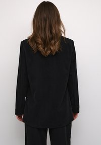 Blazer de pana negra con mangas largas, una silueta recta y una textura suave. Patrón acanalado visible a lo largo de la tela.