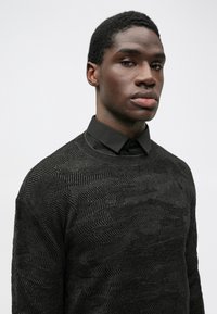 Pulover negru texturat cu un model geometric, purtat peste o cămașă neagră cu guler. Puloverul are gât rotund și manșete cu dungi.
